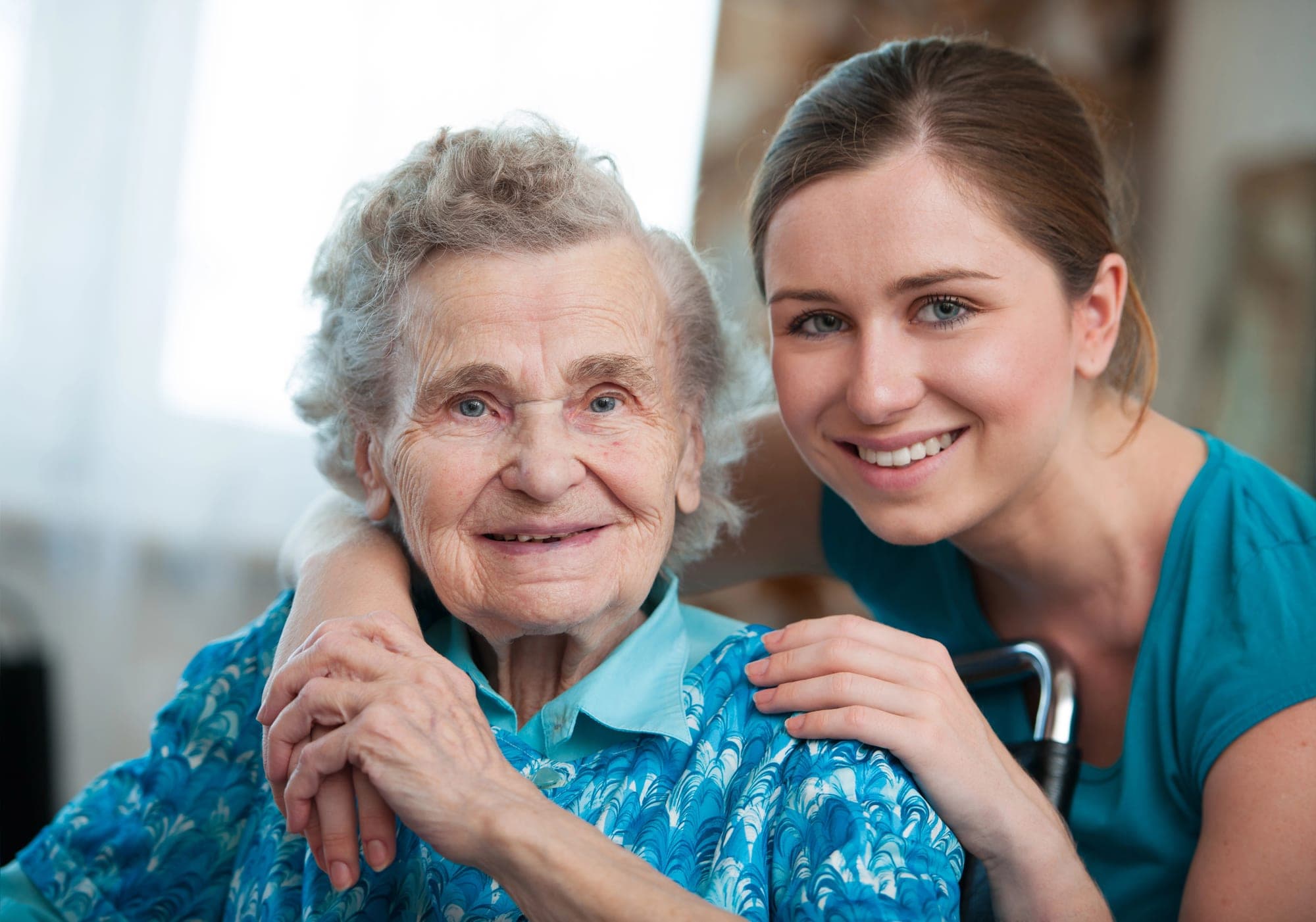 A care worker with her arm around a smiling older woman, sitting together indoors.