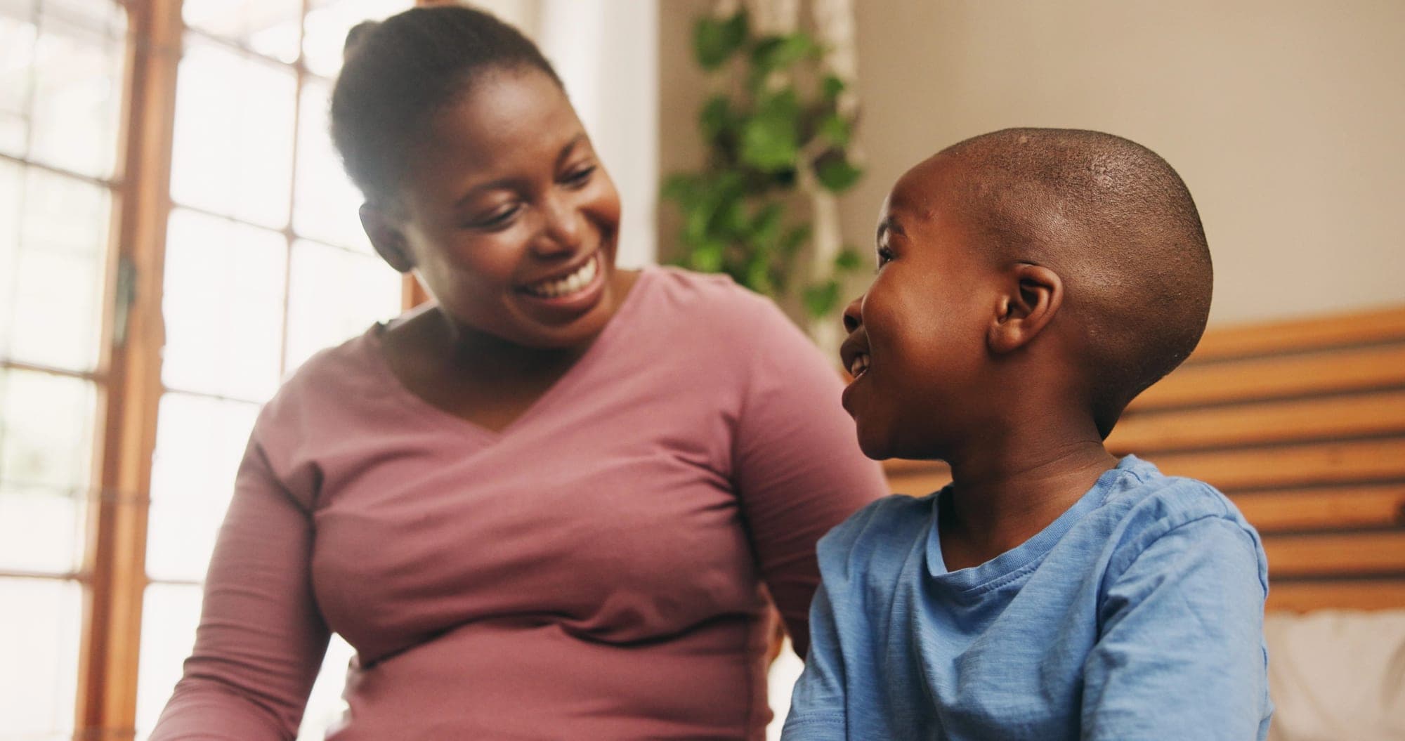 A caregiver and a young boy sitting together and laughing in a sunlit living room.